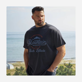 En mand står udendørs med udsigt over havet i baggrunden iført en mørk t-shirt med teksten "Mountain Expedition North Latitude" og et bjergmotiv. Han har hænderne i lommerne og kigger til siden i en afslappet positur.